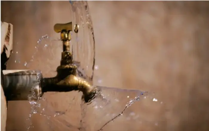 Fotografía de una llave o grifo de metal abierto, del que sale agua salpicando sobre una superficie de piedra o barro. Representa el desperdicio y la importancia de cerrar la llave.