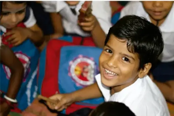 Fotografía de un niño sonriente sentado en el suelo, con otros niños alrededor, en un ambiente escolar.
