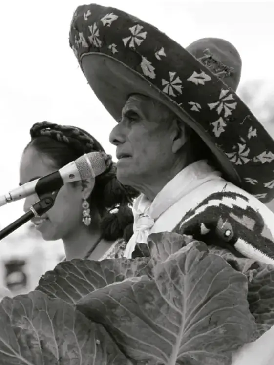 Fotografía en blanco y negro de una mujer con sombrero tradicional bordado hablando frente a un micrófono, con hojas de planta en primer plano; representa una ceremonia o evento cultural de los na savi.