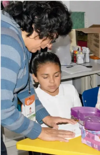 Fotografía de una mujer adulta que ayuda a una niña con uniforme blanco a trabajar en una mesa amarilla dentro del aula; representa el acompañamiento adulto en el registro de información.