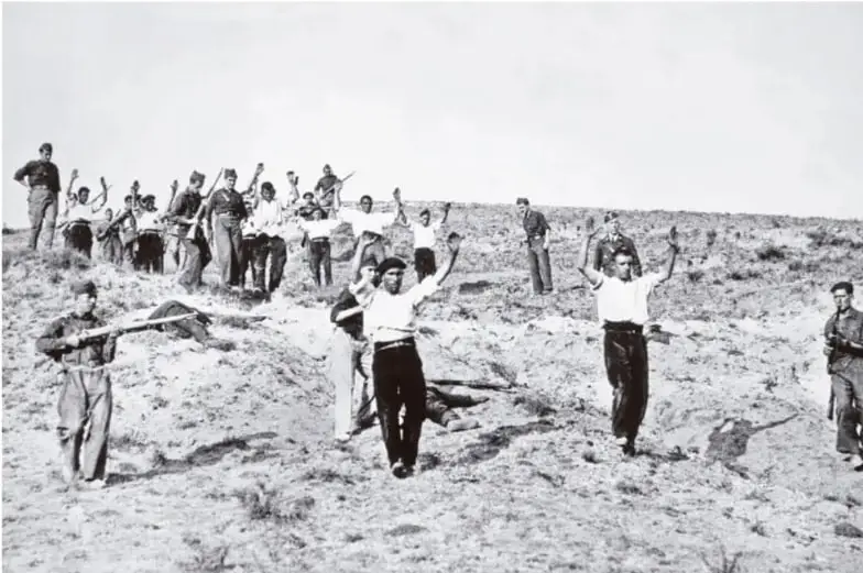 Fotografía en blanco y negro que muestra a un grupo de hombres con los brazos en alto, en un paisaje árido, custodiados por personas armadas. Representa la detención de prisioneros durante la Guerra Civil o la posguerra.