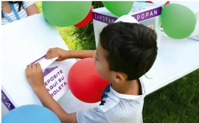 Niño depositando un voto en una urna con globos de colores.