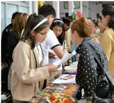 Fotografía de estudiantes atendiendo una mesa de exposición con materiales y visitantes en la galería escolar.