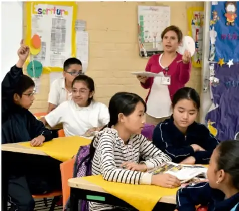 Fotografía de un salón de clases con una maestra de pie y varios estudiantes de primaria sentados en mesas amarillas, participando en una actividad grupal.