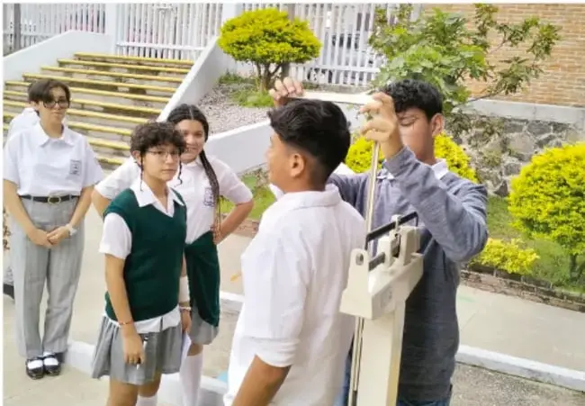 Fotografía de estudiantes de telesecundaria en el patio de su escuela, algunos en uniforme con chaleco verde, observando una medición en una balanza estadiómetro operada por un adulto.