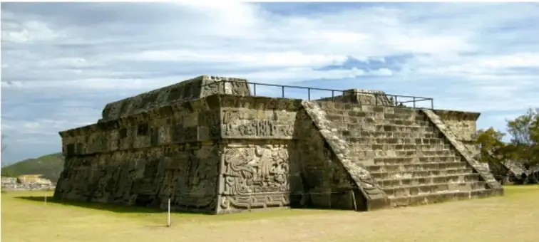 Fotografía de una plataforma o basamento de piedra mesoamericano con escalinatas y relieves tallados, rodeada de pasto y vegetación bajo un cielo con nubes (sitio arqueológico).