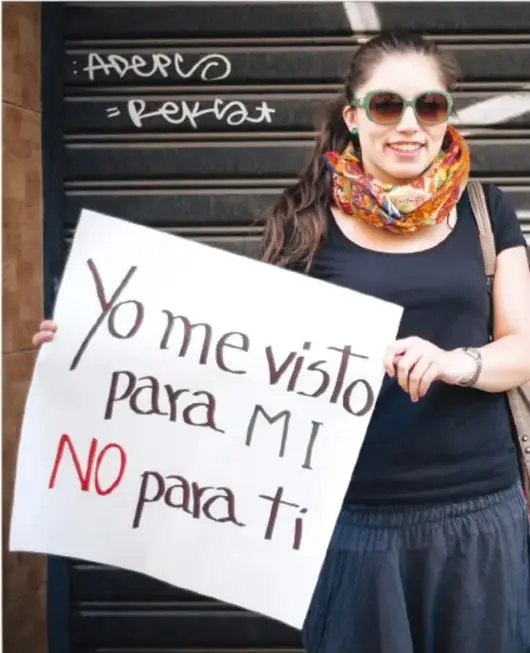 Fotografía de una mujer joven con lentes de sol y pañuelo de colores al cuello que sostiene un cartel blanco con la frase «Yo me visto para MÍ, NO para ti», en un contexto de protesta contra la violencia de género.