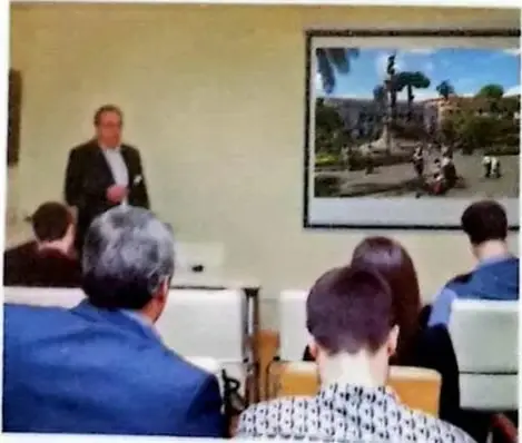 Fotografía de una persona realizando una exposición ante un público, mientras se proyecta un video en una pantalla.