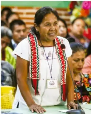 Mujer indígena sonriente con blusa blanca bordada y listones rojos habla en un micrófono durante una asamblea comunitaria, con más personas sentadas al fondo.