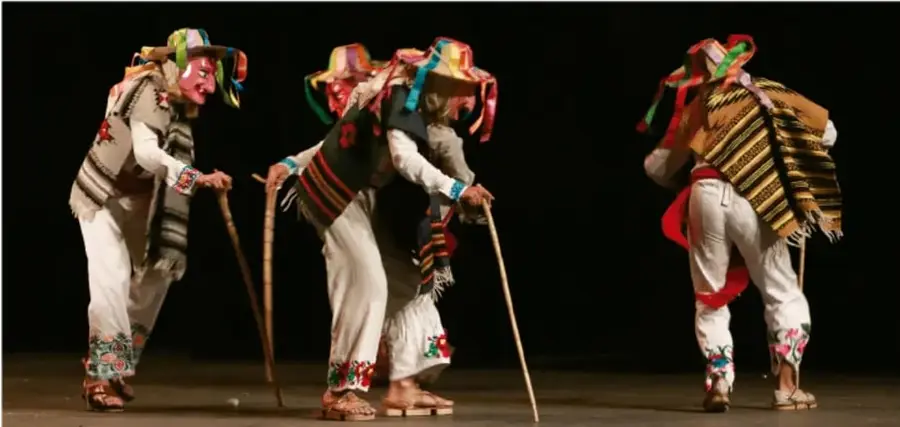 Tres danzantes con vestuario tradicional, máscaras y bastones, en un escenario oscuro; representan una danza comunitaria.