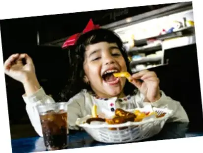 Fotografía de una niña comiendo frituras y bebiendo refresco en un establecimiento de comida rápida.