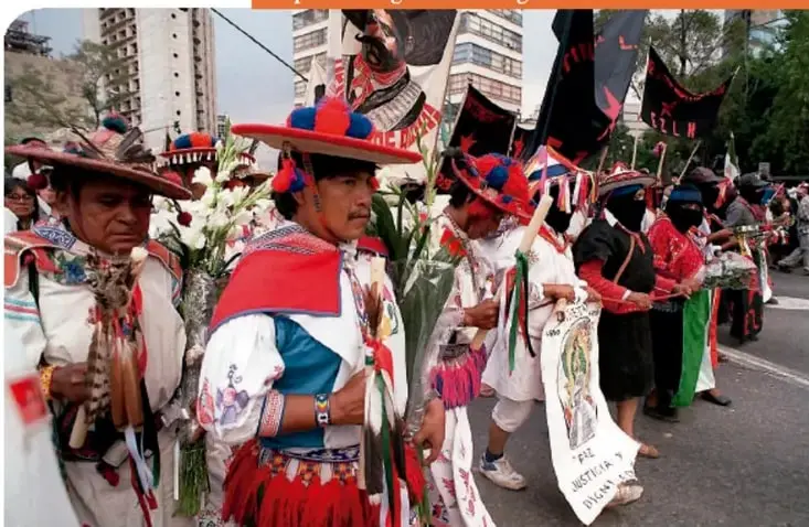 Fotografía a color de la Caravana por la Dignidad Indígena en la Ciudad de México, con personas con vestimenta tradicional y banderas, con la leyenda: 'Llegada a la Ciudad de México de la Caravana por la Dignidad Indígena en marzo de 2001'.