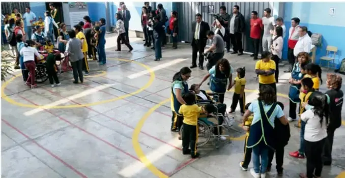 Fotografía de un simulacro escolar en un patio techado: estudiantes y personal reunidos, algunos con cascos amarillos y chalecos, atendiendo a una persona en silla de ruedas como parte del ejercicio de evacuación y rescate.