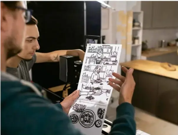 Fotografía de dos personas revisando una página impresa con viñetas de historieta dibujada a mano, en un estudio o taller creativo.
