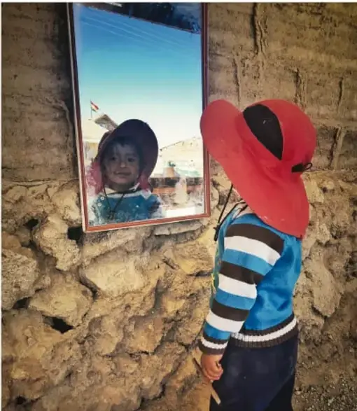 Fotografía de un niño con un sombrero rojo grande observándose en un espejo colgado sobre una pared de piedra; se refleja su rostro sonriente.