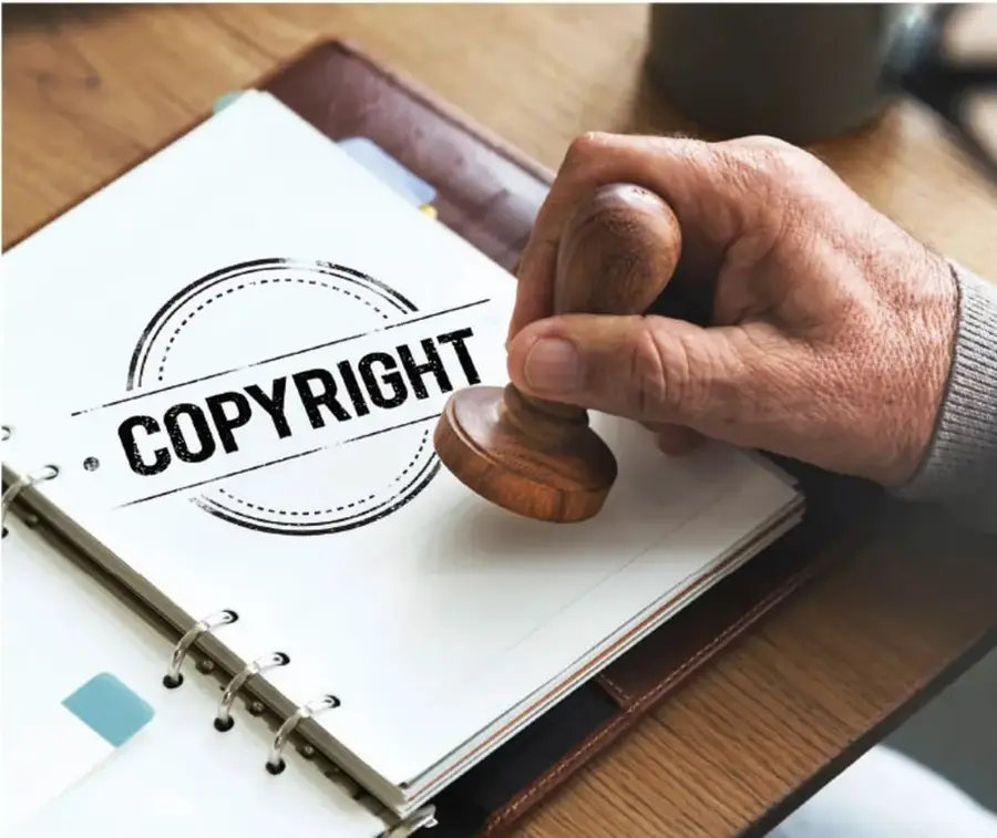 Fotografía de una mano sosteniendo un sello de madera con la palabra 'COPYRIGHT' sobre un cuaderno de argollas abierto, sobre una mesa de madera. Simboliza la propiedad intelectual y los derechos de autor.