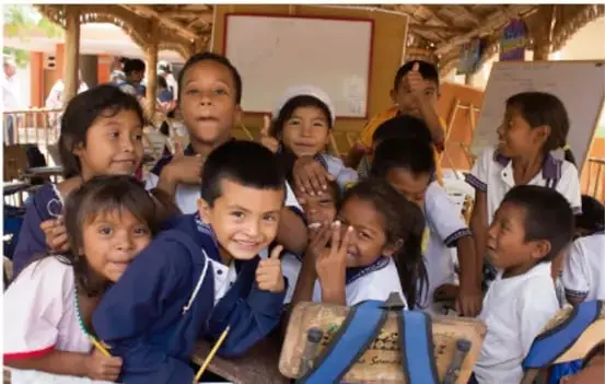 Estudiantes de primaria sentados en un aula rural, sonriendo y levantando la mano.