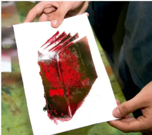 Fotografía de un estudiante sosteniendo con ambas manos una obra de arte impresa: una impresión o monoprint con manchas de pintura roja, verde y marrón sobre un papel blanco rectangular; se aprecian las manos del estudiante y parte de su torso.