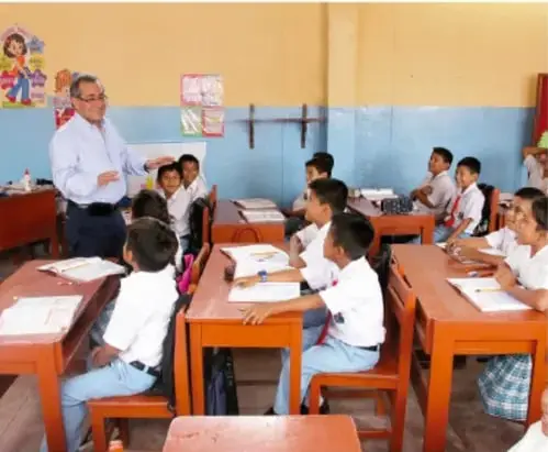 Fotografía de un aula con un maestro y estudiantes de primaria trabajando en sus pupitres.