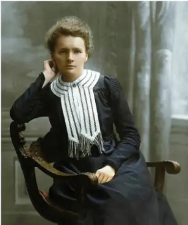 Retrato fotográfico en blanco y negro de Marie Curie, sentada en un sillón, con vestido oscuro y cuello blanco bordado.
