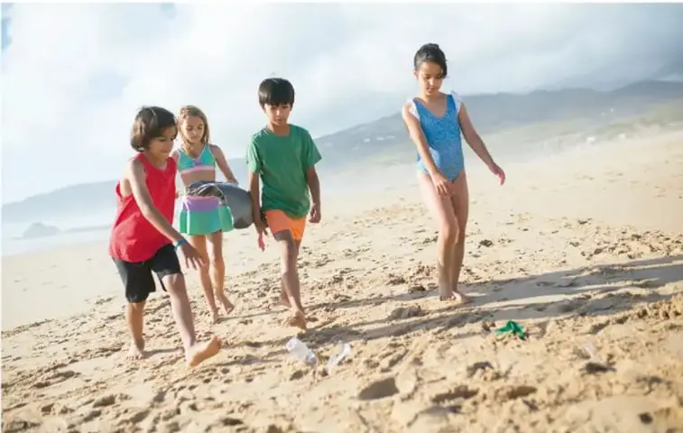 Cuatro niños y niñas en una playa recogiendo residuos; uno de ellos lleva una bolsa, en un entorno de limpieza ambiental.