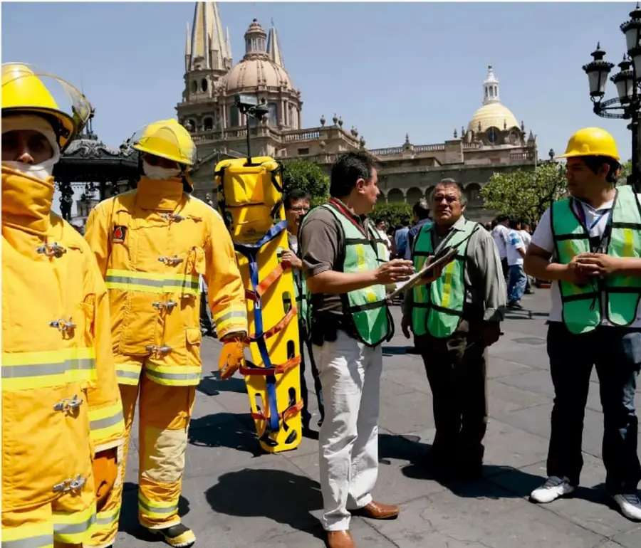 Fotografía de personal de protección civil: dos personas con trajes amarillos de protección y cascos, un operador con mochila amarilla de equipo de rescate, y tres personas con chalecos verdes de coordinación (una con clipboard), en una plaza pública con edificios históricos (catedral y domo) al fondo.
