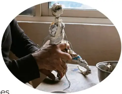 Fotografía en óvalo del artesano sosteniendo la figura de papel en proceso (un pequeño ser con cabeza) mientras sigue forrándola con engrudo y papel.