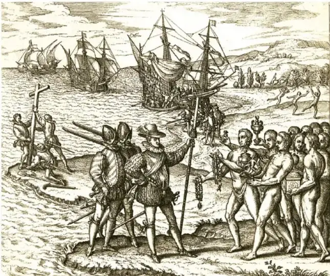 Grabado histórico que muestra la llegada de Colón a América. Se observan barcos europeos en la costa, un grupo de españoles con armaduras y una cruz, y un grupo de indígenas desnudos que les ofrecen objetos. Al fondo, más naves y personas.