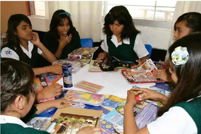 Fotografía de seis estudiantes de secundaria con uniforme, sentados alrededor de una mesa trabajando en equipo con recortes, revistas, colores y su álbum de proyecto de inglés.