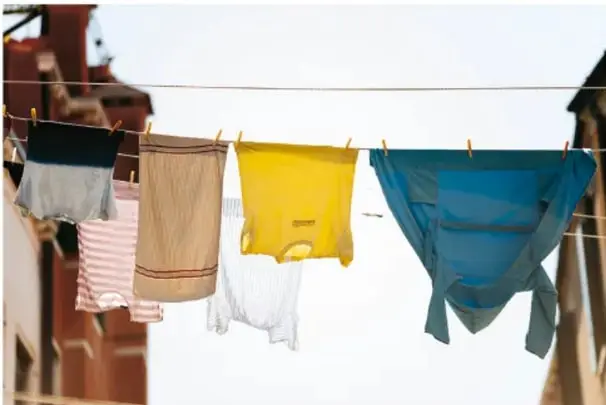 Fotografía de un tendedero con ropa de distintos colores (gris, rayas rosas, beige, amarillo y azul) secándose al sol, con edificios de fondo. Ilustra el uso cotidiano y directo de la energía solar.