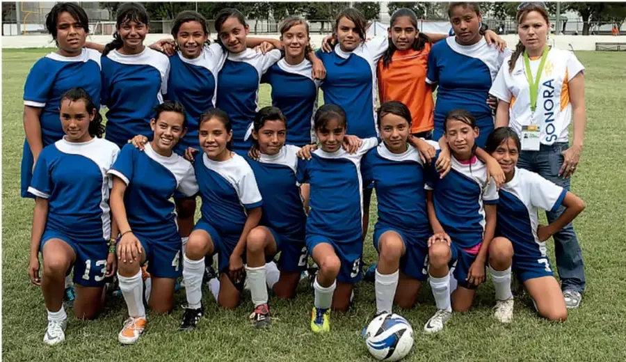 Fotografía de un equipo juvenil femenil de fútbol con uniforme azul y blanco, junto a su entrenadora, en una cancha de pasto; una futbolista con chamarra naranja (portera) y una pelota en el suelo.