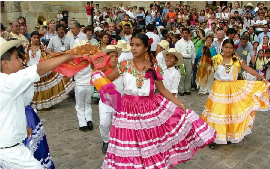Fotografía de una plaza donde niñas y niños bailan con faldas amplias coloridas (rosa y amarilla) y camisas blancas, con sombreros, ante una multitud de espectadores; representa una celebración tradicional comunitaria en México.