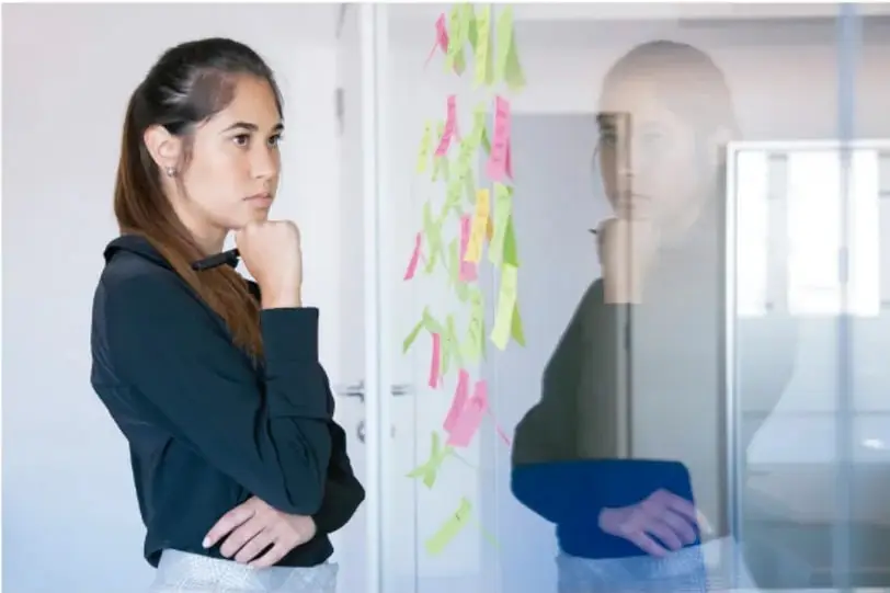Fotografía de una joven pensativa con la mano en el mentón, de pie frente a un ventanal o muro de vidrio cubierto con notas adhesivas de colores (verde, rosa, amarillo). Se refleja su rostro en el vidrio. La imagen ilustra el proceso de reflexión y organización de ideas para tomar decisiones.