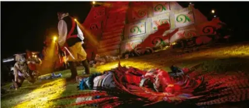 Fotografía de una presentación artística nocturna al aire libre: escenario iluminado con telones rojos y figuras, público o integrantes recostados en el suelo, ambiente de evento cultural comunitario.