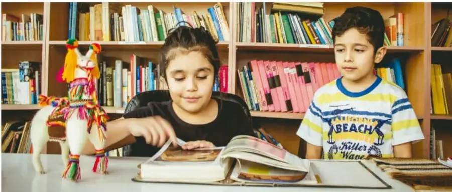 Fotografía de dos niños en una biblioteca escolar. Una niña hojea un libro abierto mientras un niño la observa. Sobre la mesa hay un libro abierto y un caballito artesanal de tela con listones de colores. Detrás se ven estantes llenos de libros.