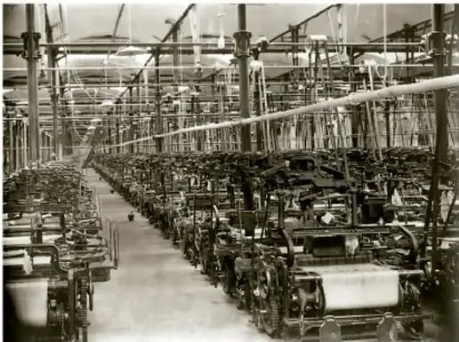 Fotografía histórica en blanco y negro: interior de una hilandería con largas hileras de máquinas textiles accionadas por transmisiones de correa, representando la producción industrial.