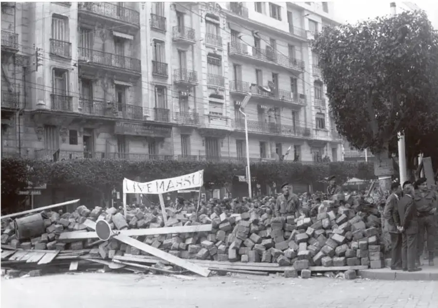 Fotografía en blanco y negro de una calle de Argel con una gran barricada de escombros y ladrillos. Se ve un cartel con la palabra 'LIVEMESS' y varias personas, algunas con uniforme militar, junto a la barricada. Al fondo, edificios de varios pisos con balcones.