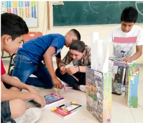 Fotografía de estudiantes de telesecundaria trabajando en equipo en el piso del salón con materiales impresos, libros y una maqueta o proyecto elaborado; créditos: Jesús Eduardo Arteaga Flores, Tamaulipas.
