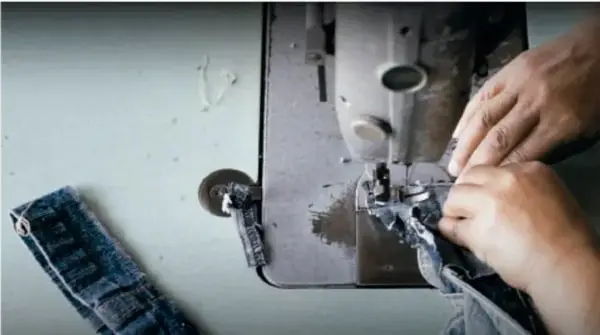 Fotografía de manos cosiendo tela con una máquina de coser industrial; se observa una prenda de mezclilla y herramientas de costura sobre una mesa metálica.