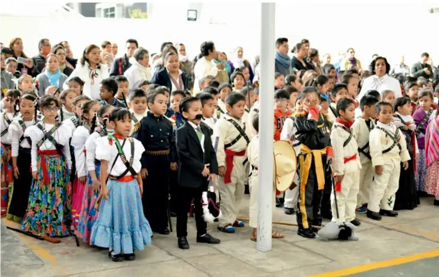Fotografía de un grupo numeroso de niños y niñas de primaria vestidos con trajes típicos y ropa de gala, acompañados por sus familias, reunidos en el patio de una escuela durante una celebración comunitaria.