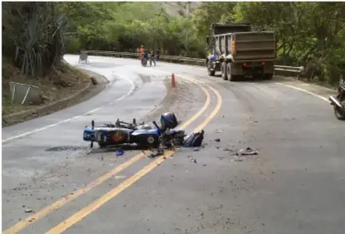 Fotografía de un accidente de tránsito en una carretera con curva: una motocicleta caída en el pavimento, personal de emergencias con chalecos reflectivos y un camión de carga detenido al costado.