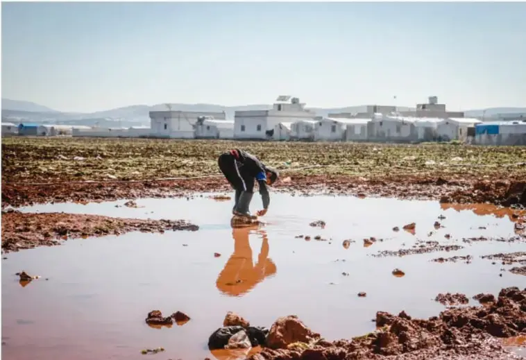 Fotografía de una charca de agua rojiza en una zona árida con residuos mineros; una persona se agacha junto al agua, con edificios blancos de fondo, ilustrando la contaminación por hierro y metales pesados.