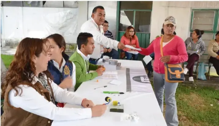 Mesa de casilla electoral al aire libre: funcionarios de casilla entregan una papeleta a una votante; otras personas esperan su turno.
