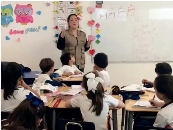 Fotografía de un aula de primaria: una maestra de pie frente al pizarrón presenta una actividad mientras varios estudiantes sentados en sus bancas la observan y toman notas. Hay decoraciones de foami y corazones en la pared.