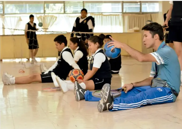 Estudiantes sentados en el piso de un gimnasio con pelotas, realizando actividad física.