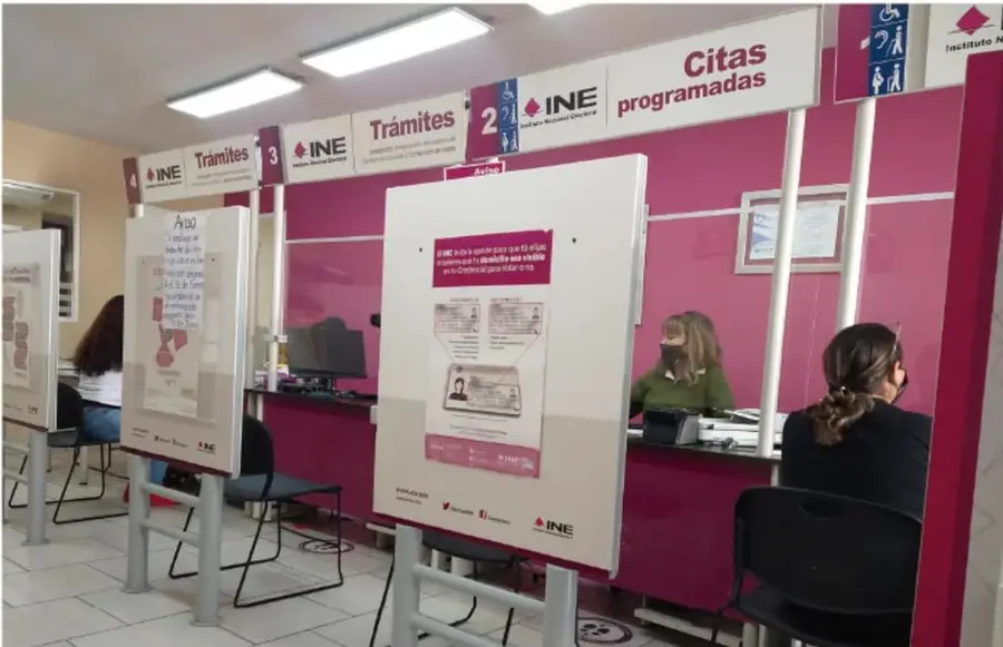 Fotografía de módulos de atención del Instituto Nacional Electoral (INE) con rótulos «Trámites» y «Citas programadas»; se observa personal atendiendo a ciudadanas tras mamparas, con carteles informativos sobre el padrón electoral.