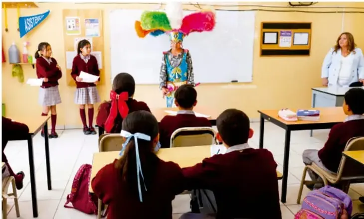 Fotografía de un aula de telesecundaria en México: estudiantes en uniforme frente al pizarrón, una persona disfrazada de artista en el centro, compañeros sentados y una maestra de pie a la derecha; parece una muestra escénica o declamación.