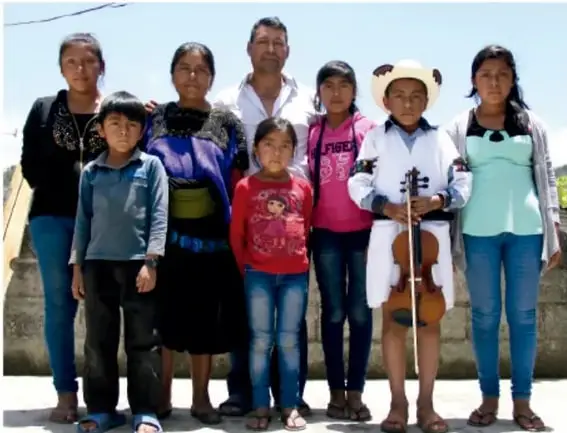 Fotografía de un grupo de personas de diversas edades, aparentemente de una comunidad originaria, posando al aire libre; una niña porta un sombrero y un violín.