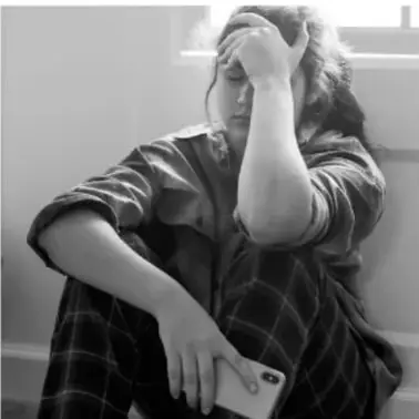 Fotografía en blanco y negro de una persona joven sentada, con la mano en la cabeza, sosteniendo un teléfono celular; expresa angustia.