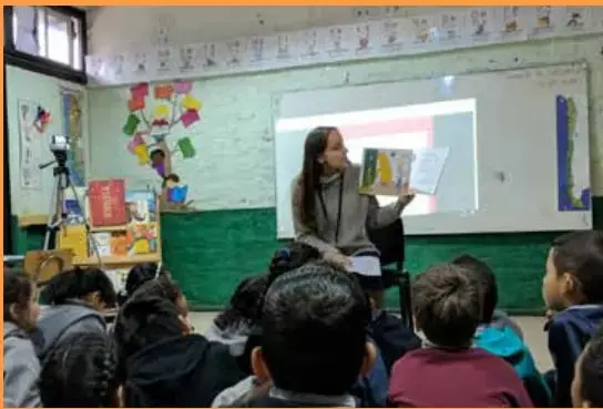 Fotografía de una sala de clases donde la docente lee un libro en voz alta frente a sus estudiantes, quienes están sentados en el suelo mirándola. Al fondo hay una pizarra y materiales educativos.